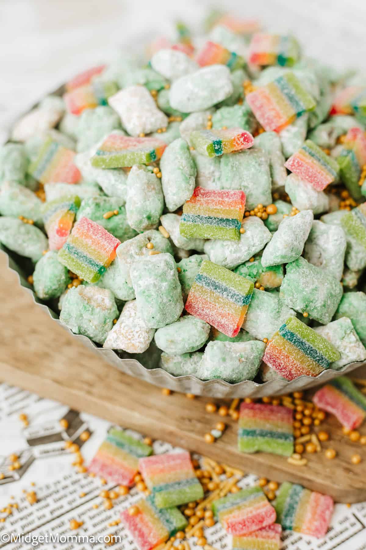 A bowl of green powdered snacks mixed with colorful striped candy pieces and gold sprinkles, placed on a wooden board and newspaper.