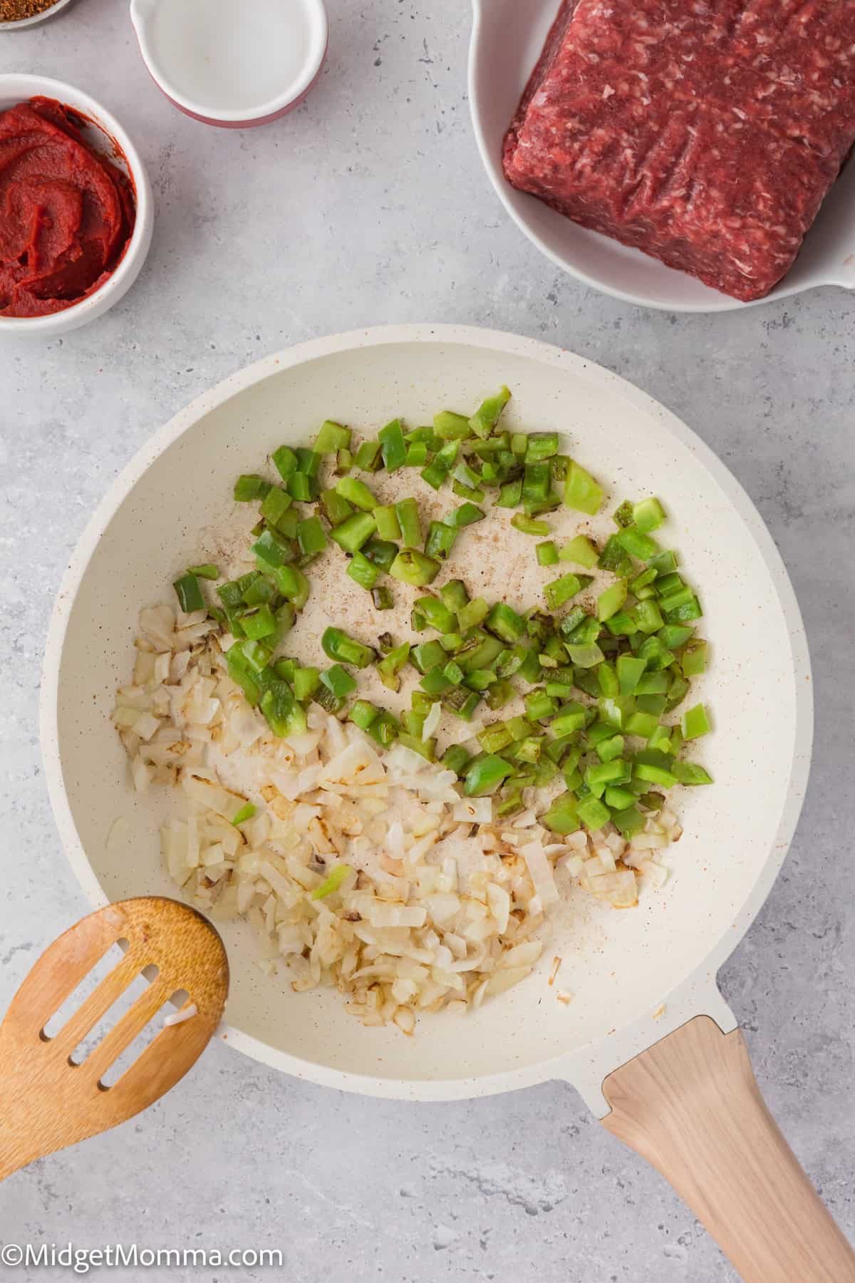 Chopped onions and green peppers cooking in a white pan, with a wooden spatula. Ground beef and tomato paste are nearby in bowls.