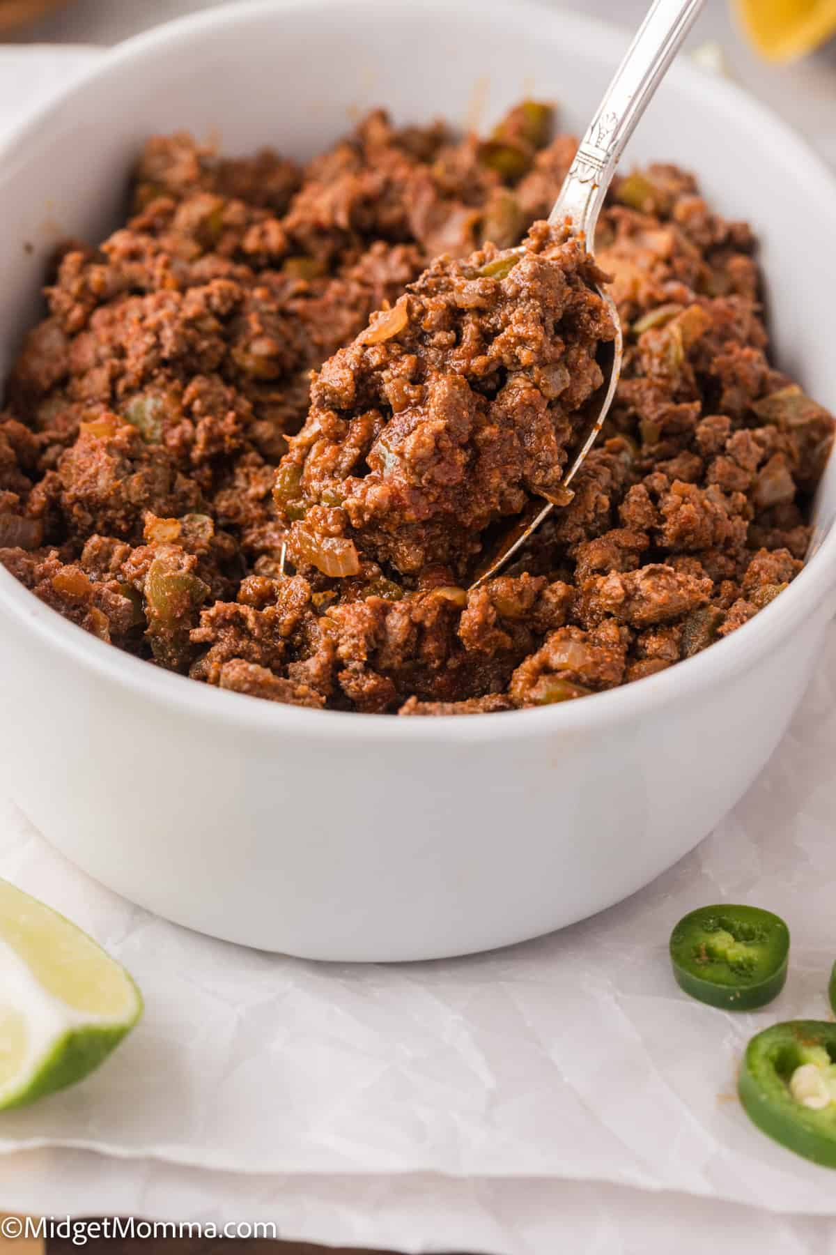 A bowl of seasoned ground beef with green peppers, showing a spoonful. There are sliced jalapeños and a lime wedge nearby.