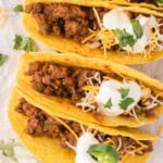 Three hard-shell tacos filled with seasoned ground meat, topped with shredded cheese, sour cream, and sliced jalapeños, garnished with cilantro.