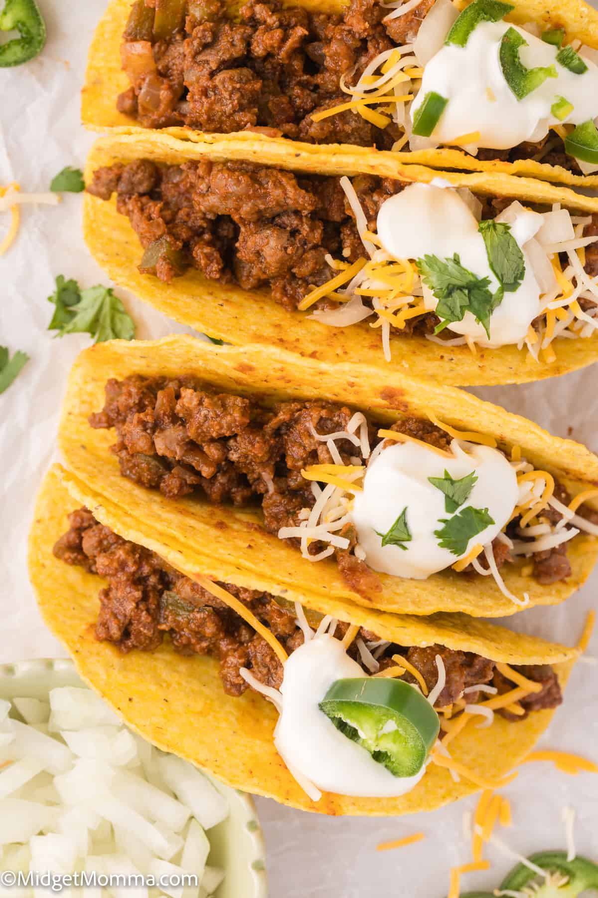 Three hard-shell tacos filled with seasoned ground meat, topped with shredded cheese, sour cream, and sliced jalapeños, garnished with cilantro.