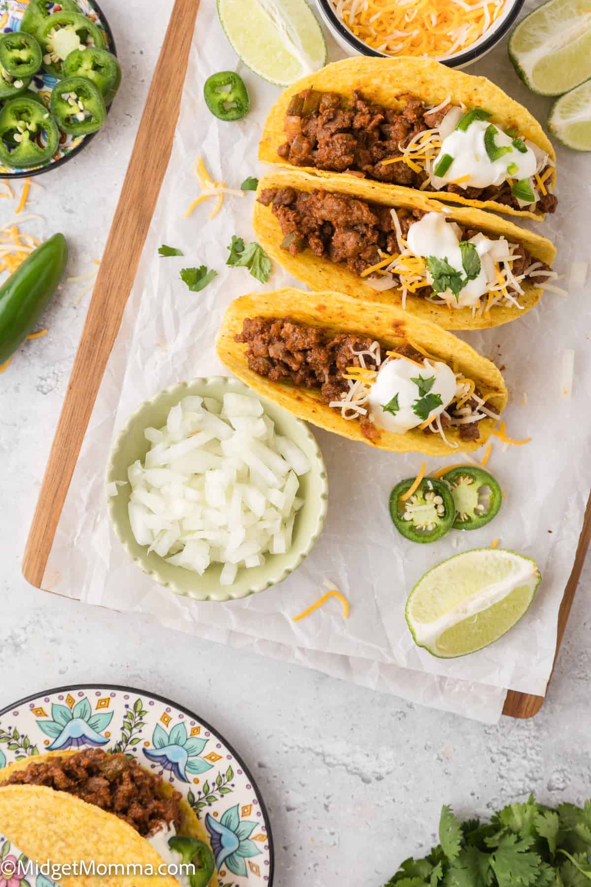 Three tacos with ground meat, cheese, and sour cream on a wooden board, surrounded by lime wedges, sliced jalapeños, and a bowl of chopped onions.