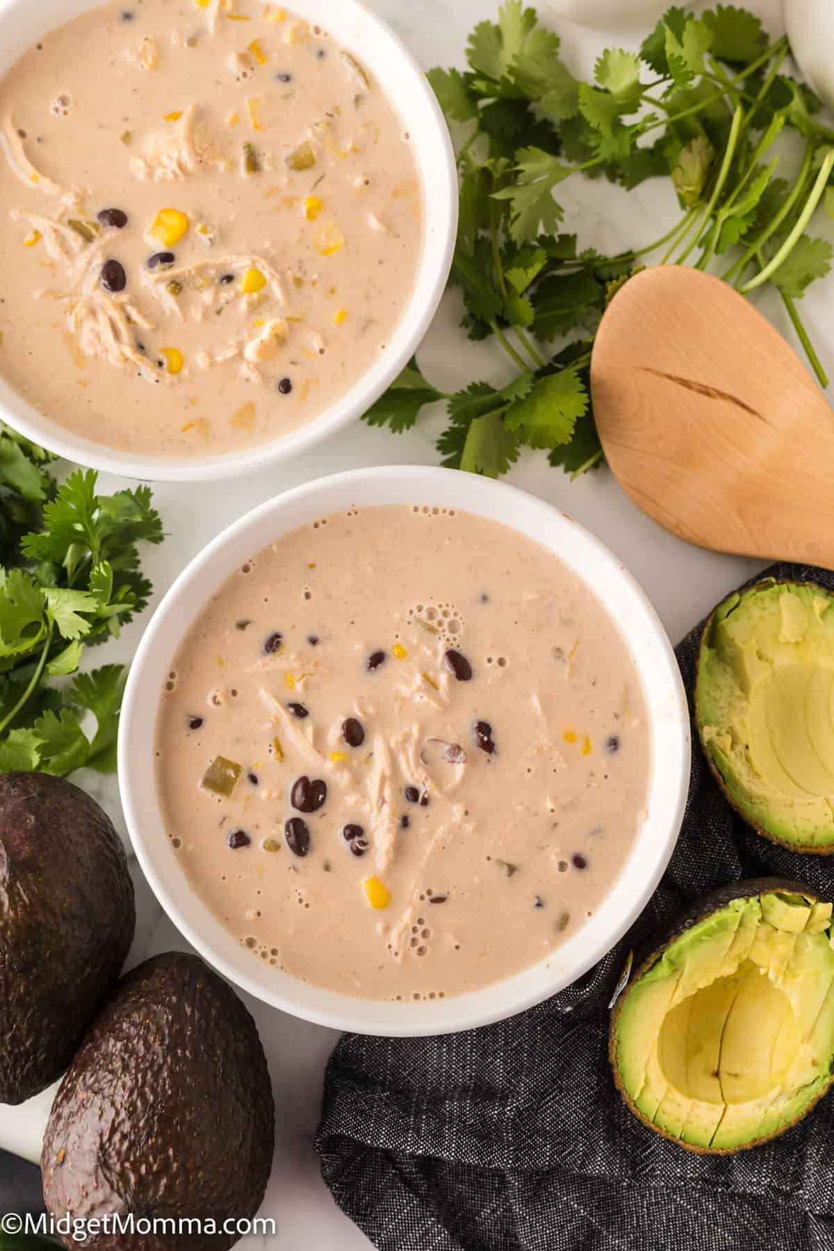 Two bowls of creamy white chicken chili with corn and black beans, surrounded by avocados, cilantro, and a wooden spoon.