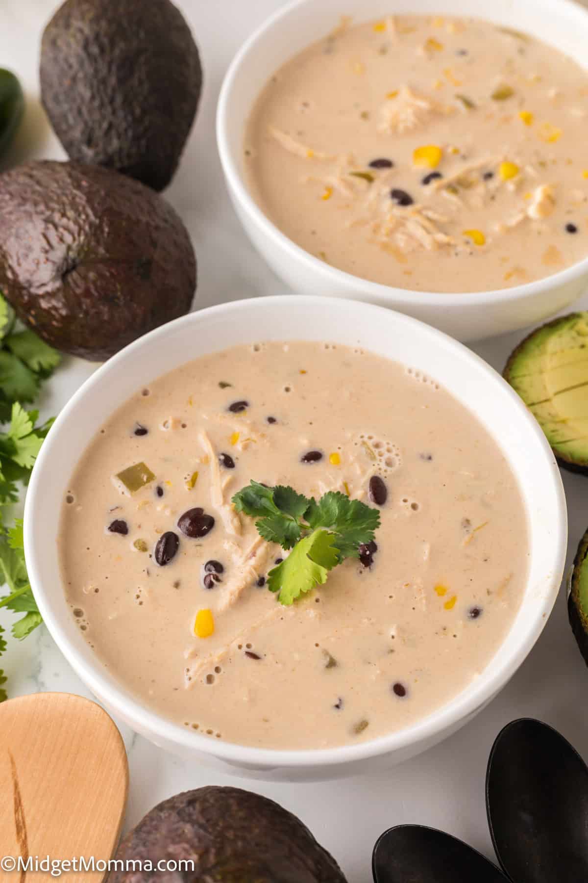 Two bowls of creamy soup with black beans, corn, and chicken, garnished with cilantro. Surrounding are avocados and a wooden spoon.