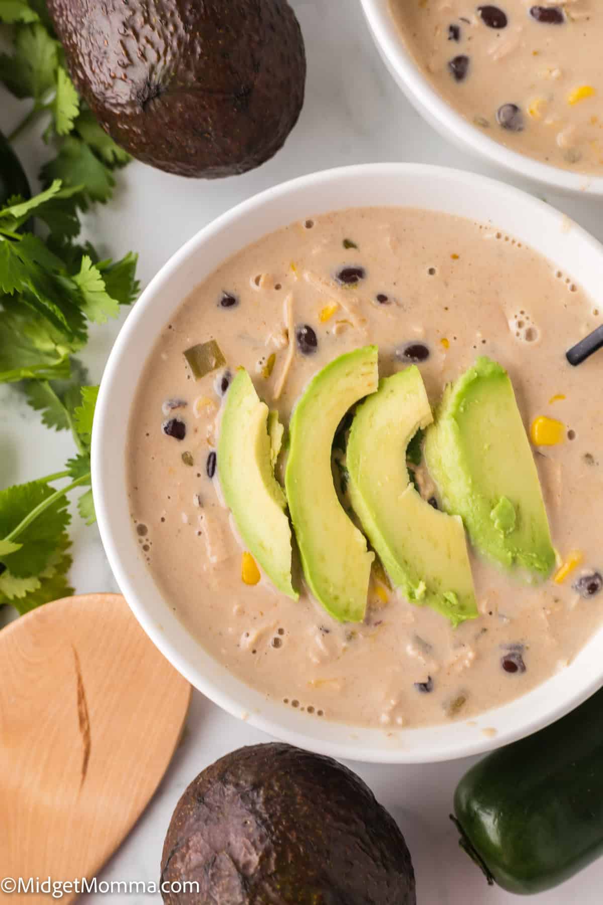 Creamy white chicken chili in a bowl topped with sliced avocado. Surrounding ingredients include avocados, cilantro, and a jalapeño pepper.