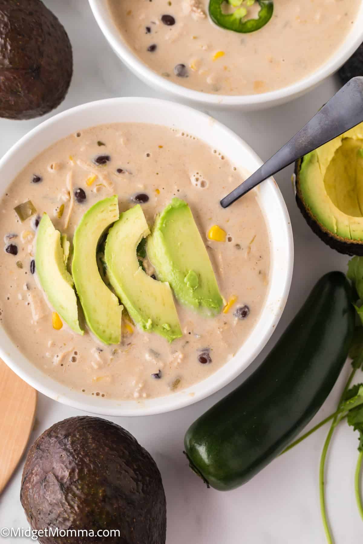 A bowl of creamy white chicken chili with black beans, corn, and avocado slices. A spoon rests in the bowl. Avocado halves and a jalapeño pepper are nearby.