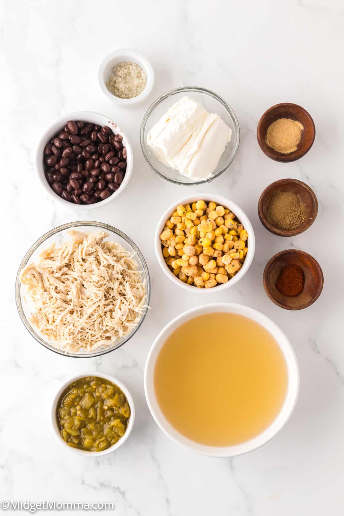 white chicken chili ingredients. Various ingredients, including black beans, chickpeas, shredded cheese, green chilies, and spices, are arranged in bowls on a white surface.